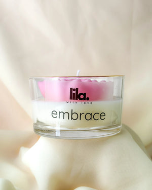 Candle in a glass jar with 'lila. with love' branding and 'embrace' text on a soft background.