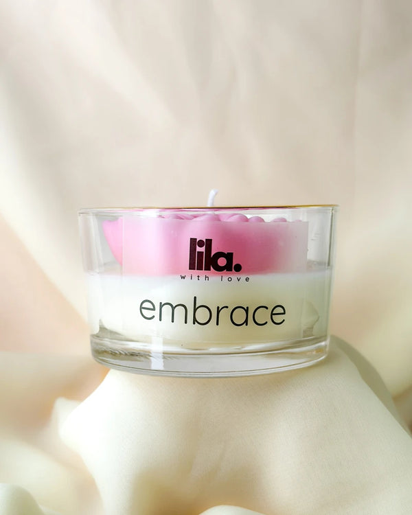 Candle in a glass jar with 'lila. with love' branding and 'embrace' text on a soft background.