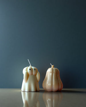 Two unique candle designs, one shaped like a ghost and the other like a pumpkin, on a reflective surface with a dark background.