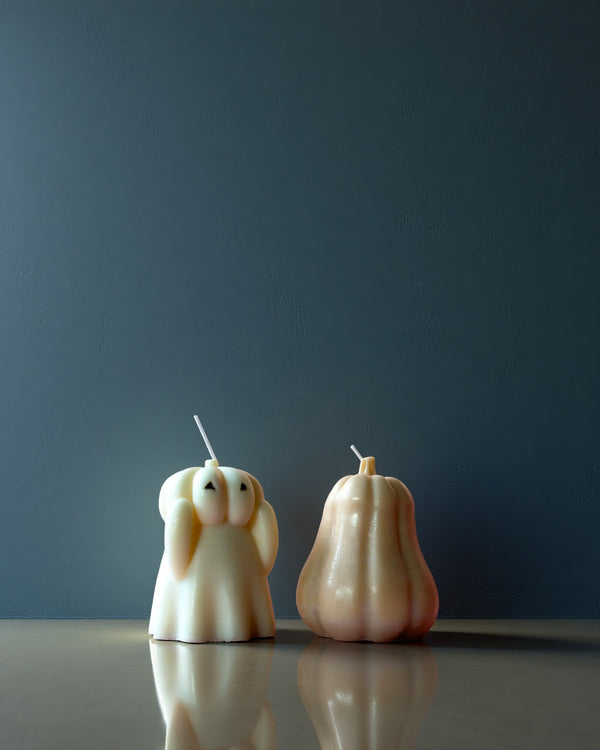 Two unique candle designs, one shaped like a ghost and the other like a pumpkin, on a reflective surface with a dark background.