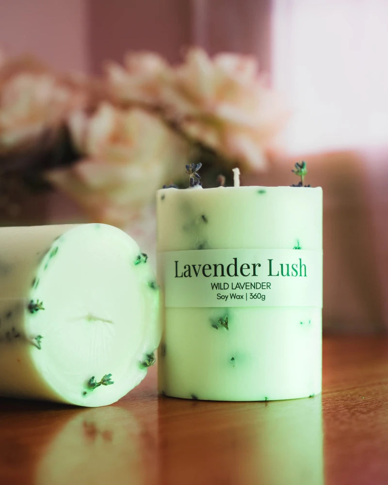 'Lavender Lush' pillar candle from Lila with love on a wooden surface with blurred flowers in the background.