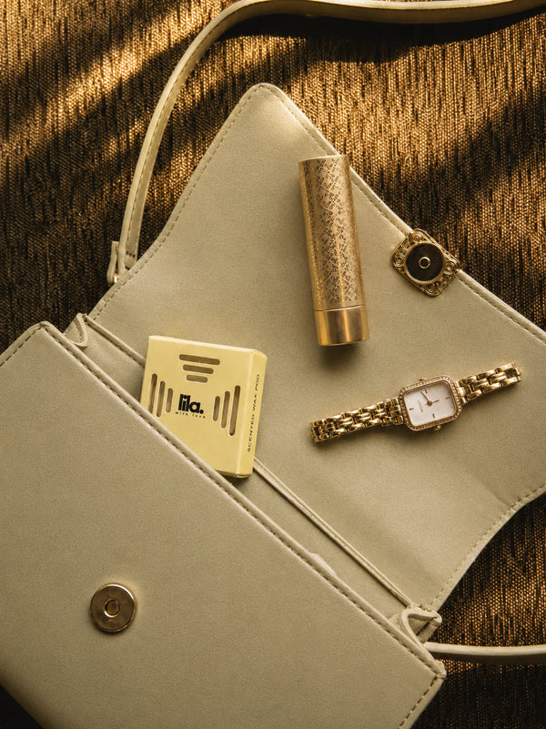 A yellow lila with love scented wax pod tucked inside a cream evening clutch beside a gold lipstick, watch and earring