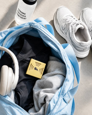 A yellow lila with love scented wax pod tucked into a blue gym bag with workout clothes and headphones, beside trainers and a water bottle