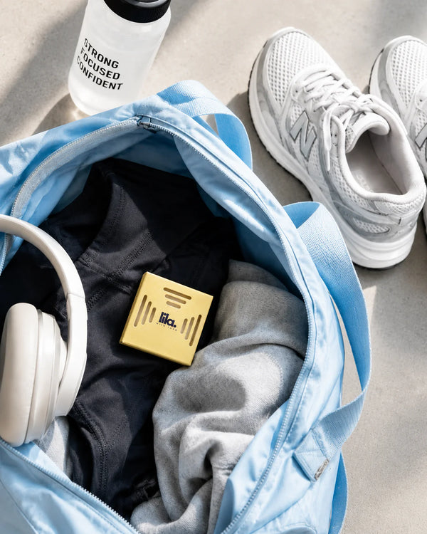 A yellow lila with love scented wax pod tucked into a blue gym bag with workout clothes and headphones, beside trainers and a water bottle