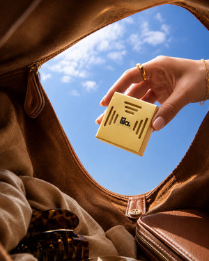 A hand drops a yellow lila with love scented wax pod into a tan suede handbag, seen from inside the bag against a blue sky