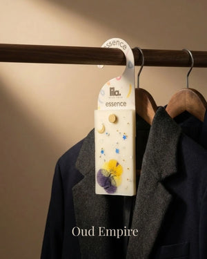 wax sachet from lila with love with floral designs and golden beads  hanging on a wooden hanger on wooden wardrobe with cloths around them