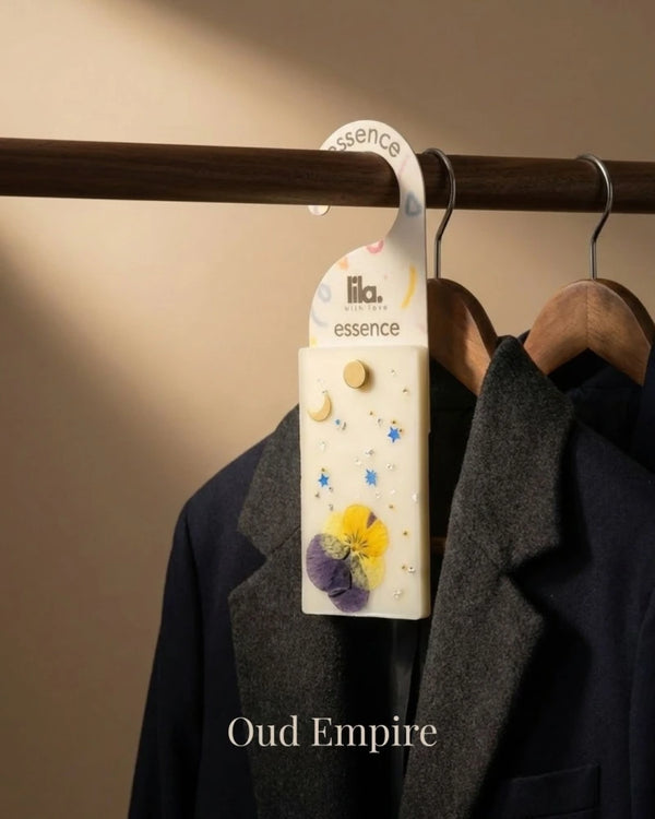 wax sachet from lila with love with floral designs and golden beads  hanging on a wooden hanger on wooden wardrobe with cloths around them