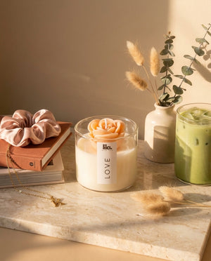 Candle with rose-shaped wax in a glass jar on a marble surface with books and decorative items.