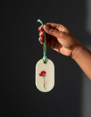 Hand holding a white wax sachet with red flower  and green stem against a dark background